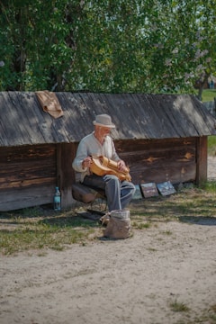 An elderly man sits on a stool playing a stringed musical instrument in a rustic outdoor setting. He wears a light-colored hat and shirt. Behind him, a wooden structure provides a backdrop with trees and greenery visible in the background. On the ground near him, there is a woven bag and some artwork displayed.