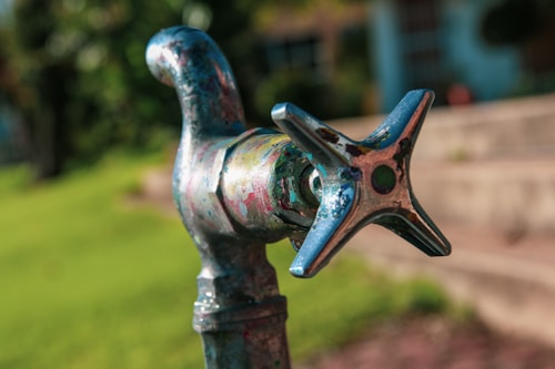 A close-up of a weathered outdoor faucet with a cross-shaped handle, featuring multicolored patches of paint or rust. The background is blurred, with hints of greenery and outdoor steps, suggesting a garden or yard setting.
