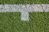 A patch of artificial grass with a thick white line running horizontally across it, intersected by a shorter vertical white line.