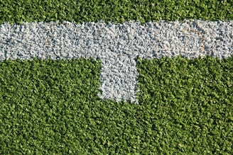 A patch of artificial grass with a thick white line running horizontally across it, intersected by a shorter vertical white line.