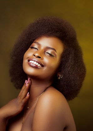 A woman with a radiant smile and closed eyes. Her hand is gently touching her neck. She has short, curly hair and is wearing subtle eye makeup. The background is a warm, soft yellow-brown tone.