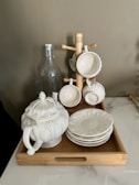 A wooden tray holds a porcelain teapot with ornate designs, several matching saucers, and a wooden stand supporting four white teacups. A tall, clear glass bottle with a stopper sits nearby.