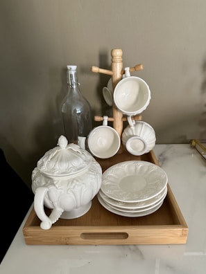 A wooden tray holds a porcelain teapot with ornate designs, several matching saucers, and a wooden stand supporting four white teacups. A tall, clear glass bottle with a stopper sits nearby.