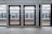 Glass platform doors at a train station with visible tracks and another platform on the opposite side. The modern structure includes metal framing and clear panels, providing a view of the train tracks and reflections of station surroundings.
