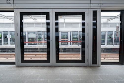 Glass platform doors at a train station with visible tracks and another platform on the opposite side. The modern structure includes metal framing and clear panels, providing a view of the train tracks and reflections of station surroundings.