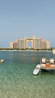 A grand, luxury hotel building with distinctive architecture stands prominently across the water. The structure features a central arch and towers with a pinkish hue. In the foreground, a small dock with seats and the clear blue ocean can be seen, adding a serene and opulent atmosphere to the scene.