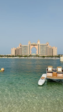 A grand, luxury hotel building with distinctive architecture stands prominently across the water. The structure features a central arch and towers with a pinkish hue. In the foreground, a small dock with seats and the clear blue ocean can be seen, adding a serene and opulent atmosphere to the scene.