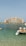 A grand, luxury hotel building with distinctive architecture stands prominently across the water. The structure features a central arch and towers with a pinkish hue. In the foreground, a small dock with seats and the clear blue ocean can be seen, adding a serene and opulent atmosphere to the scene.