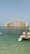 A grand, luxury hotel building with distinctive architecture stands prominently across the water. The structure features a central arch and towers with a pinkish hue. In the foreground, a small dock with seats and the clear blue ocean can be seen, adding a serene and opulent atmosphere to the scene.