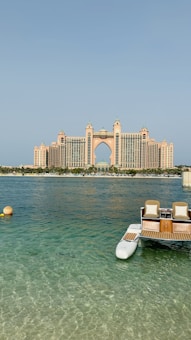 A grand, luxury hotel building with distinctive architecture stands prominently across the water. The structure features a central arch and towers with a pinkish hue. In the foreground, a small dock with seats and the clear blue ocean can be seen, adding a serene and opulent atmosphere to the scene.