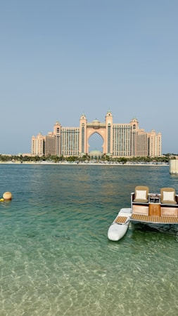 A grand, luxury hotel building with distinctive architecture stands prominently across the water. The structure features a central arch and towers with a pinkish hue. In the foreground, a small dock with seats and the clear blue ocean can be seen, adding a serene and opulent atmosphere to the scene.