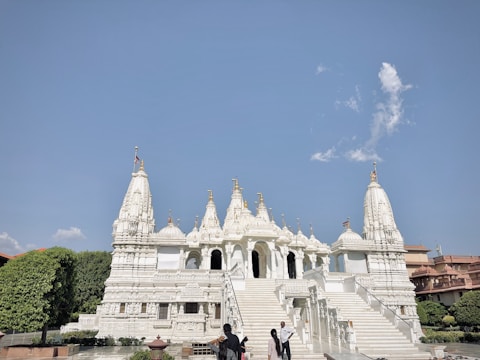 A majestic white marble temple with intricate carvings and multiple spires set against a clear blue sky. A few people are standing in the foreground near the steps leading up to the temple. Lush greenery and other architectural structures surround the area.