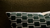 A cushioned pillow with a geometric pattern in dark green and white rests on a textured dark brown fabric. The pillow features a series of outlined rectangular shapes sewn in a consistent pattern.