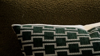 A cushioned pillow with a geometric pattern in dark green and white rests on a textured dark brown fabric. The pillow features a series of outlined rectangular shapes sewn in a consistent pattern.