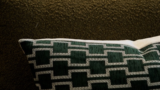A cushioned pillow with a geometric pattern in dark green and white rests on a textured dark brown fabric. The pillow features a series of outlined rectangular shapes sewn in a consistent pattern.