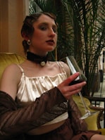 A person in vintage attire, featuring a satin top and accessories such as pearls and a choker, is seated on a green couch holding a glass of red wine. The hairstyle is retro, and the setting includes lush green plants in the background.