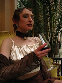 A person in vintage attire, featuring a satin top and accessories such as pearls and a choker, is seated on a green couch holding a glass of red wine. The hairstyle is retro, and the setting includes lush green plants in the background.