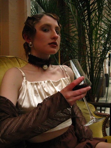 A person in vintage attire, featuring a satin top and accessories such as pearls and a choker, is seated on a green couch holding a glass of red wine. The hairstyle is retro, and the setting includes lush green plants in the background.