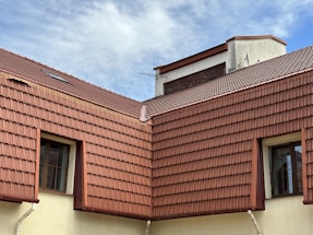 The image features a section of a building with multiple gabled roofs covered with red-brown tiles. Two windows with black frames are visible, nestled between the roof tiles and light-colored walls. A small antenna is attached to the top section of the roof, and the scene is set against a cloudy blue sky.