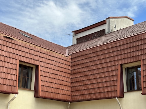 The image features a section of a building with multiple gabled roofs covered with red-brown tiles. Two windows with black frames are visible, nestled between the roof tiles and light-colored walls. A small antenna is attached to the top section of the roof, and the scene is set against a cloudy blue sky.