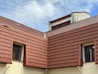 The image features a section of a building with multiple gabled roofs covered with red-brown tiles. Two windows with black frames are visible, nestled between the roof tiles and light-colored walls. A small antenna is attached to the top section of the roof, and the scene is set against a cloudy blue sky.