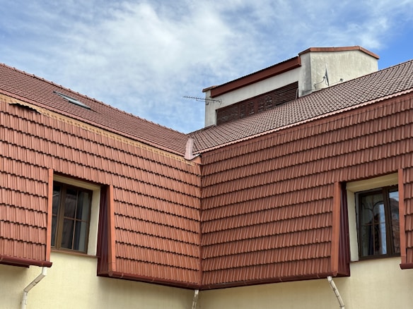The image features a section of a building with multiple gabled roofs covered with red-brown tiles. Two windows with black frames are visible, nestled between the roof tiles and light-colored walls. A small antenna is attached to the top section of the roof, and the scene is set against a cloudy blue sky.