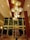 An elegant hotel lobby features a large, modern chandelier with multiple warm-colored, circular lights hanging from a high wooden ceiling. Below, there is a beautifully arranged centerpiece with tall glass vases filled with various flowers, including yellow, white, and orange blossoms. Comfortable seating is arranged throughout the lobby, and large windows provide a view to the outside.