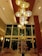 An elegant hotel lobby features a large, modern chandelier with multiple warm-colored, circular lights hanging from a high wooden ceiling. Below, there is a beautifully arranged centerpiece with tall glass vases filled with various flowers, including yellow, white, and orange blossoms. Comfortable seating is arranged throughout the lobby, and large windows provide a view to the outside.