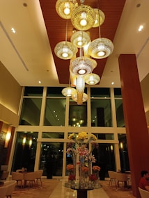 An elegant hotel lobby features a large, modern chandelier with multiple warm-colored, circular lights hanging from a high wooden ceiling. Below, there is a beautifully arranged centerpiece with tall glass vases filled with various flowers, including yellow, white, and orange blossoms. Comfortable seating is arranged throughout the lobby, and large windows provide a view to the outside.