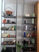 A well-organized shelf unit against a white brick wall contains various decorative items, including wicker baskets, metal pots, colorful glass bottles, small potted plants, and storage boxes. A green plant is positioned in front of the shelves.