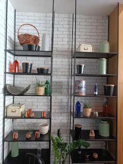 A well-organized shelf unit against a white brick wall contains various decorative items, including wicker baskets, metal pots, colorful glass bottles, small potted plants, and storage boxes. A green plant is positioned in front of the shelves.