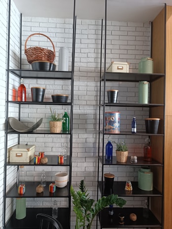 A well-organized shelf unit against a white brick wall contains various decorative items, including wicker baskets, metal pots, colorful glass bottles, small potted plants, and storage boxes. A green plant is positioned in front of the shelves.