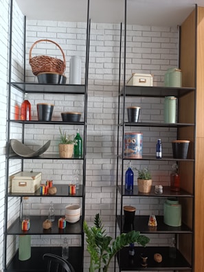 A well-organized shelf unit against a white brick wall contains various decorative items, including wicker baskets, metal pots, colorful glass bottles, small potted plants, and storage boxes. A green plant is positioned in front of the shelves.