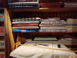 Several neatly folded fabrics in different patterns and colors are arranged on wooden shelves. Below the fabrics, there is a table with a few sewing items including a pair of scissors, a measuring tape, and a spool of thread. The shelves also have some boxes with labels. The setting gives a rustic and cozy feel.