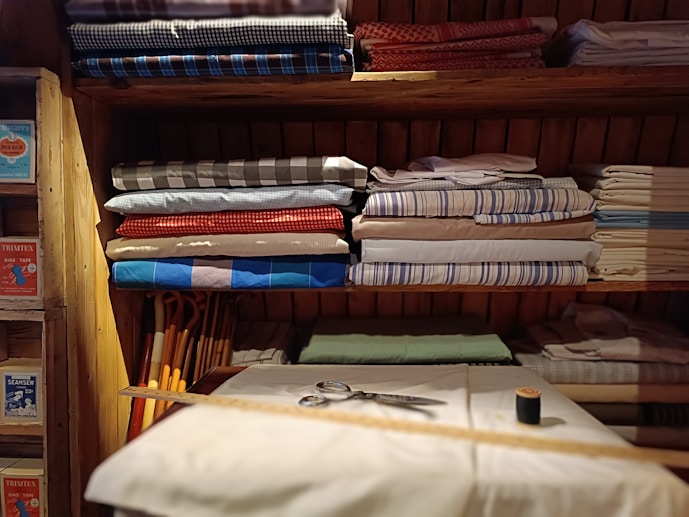 Several neatly folded fabrics in different patterns and colors are arranged on wooden shelves. Below the fabrics, there is a table with a few sewing items including a pair of scissors, a measuring tape, and a spool of thread. The shelves also have some boxes with labels. The setting gives a rustic and cozy feel.