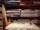 Several neatly folded fabrics in different patterns and colors are arranged on wooden shelves. Below the fabrics, there is a table with a few sewing items including a pair of scissors, a measuring tape, and a spool of thread. The shelves also have some boxes with labels. The setting gives a rustic and cozy feel.