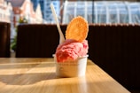 A cup of pink gelato sits on a wooden table, topped with a waffle disk and a plastic spoon. The background features a blurred view of urban buildings and large windows, suggesting an indoor location with natural light coming through.