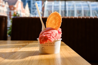 A cup of pink gelato sits on a wooden table, topped with a waffle disk and a plastic spoon. The background features a blurred view of urban buildings and large windows, suggesting an indoor location with natural light coming through.