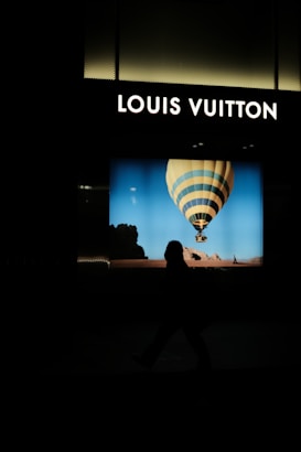 A brightly lit storefront displays the Louis Vuitton logo above an image of a hot air balloon set against a blue sky. The silhouette of a person walking by is visible in the foreground.