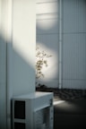 A white air conditioning unit is placed outside against a wall which is partially in shadow. Some branches with leaves are visible emerging from behind the wall. The background features a modern facade with vertical piping and a wire mesh design.