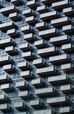 A repetitive pattern of balconies juts out from a modern high-rise building. Each balcony is made of transparent glass railings, adding a sense of uniformity and structure. The building's facade is composed of reflective glass windows, creating geometric patterns under a clear sky.