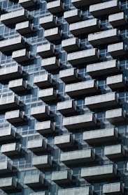 A repetitive pattern of balconies juts out from a modern high-rise building. Each balcony is made of transparent glass railings, adding a sense of uniformity and structure. The building's facade is composed of reflective glass windows, creating geometric patterns under a clear sky.