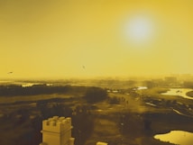 A landscape view with a vibrant golden hue, showcasing a vast area with lush greenery and water bodies. A tower-like structure is visible in the foreground, while a serene sky with a bright sun dominates the background. Birds are seen flying across the sky, adding to the scene's tranquility.