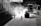 Workers are paving a road with heavy machinery in an industrial setting. The scene features silhouettes of people and vehicles backlit by the sunlight, creating a dramatic contrast between light and shadows. Equipment like a steamroller and trucks are visible.