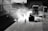 Workers are paving a road with heavy machinery in an industrial setting. The scene features silhouettes of people and vehicles backlit by the sunlight, creating a dramatic contrast between light and shadows. Equipment like a steamroller and trucks are visible.