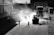Workers are paving a road with heavy machinery in an industrial setting. The scene features silhouettes of people and vehicles backlit by the sunlight, creating a dramatic contrast between light and shadows. Equipment like a steamroller and trucks are visible.