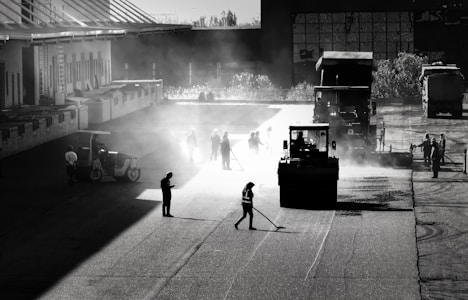 Workers are paving a road with heavy machinery in an industrial setting. The scene features silhouettes of people and vehicles backlit by the sunlight, creating a dramatic contrast between light and shadows. Equipment like a steamroller and trucks are visible.