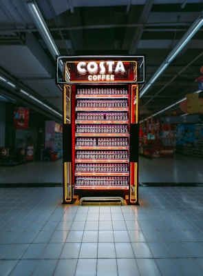 A brightly illuminated Costa Coffee fridge is filled with rows of bottled drinks, centrally placed in a large, empty aisle with a reflective tiled floor. The background is dimly lit, contrasting with the vibrant red and orange glow of the fridge.