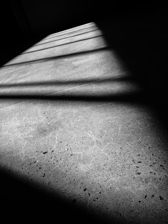 A black and white photograph of a concrete floor with a pattern of shadows cast by window blinds. The composition highlights the textures and grain of the concrete surface, intersected by linear shadow patterns that create a dynamic visual effect.