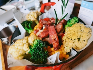A vibrant food dish featuring a mix of roasted vegetables and grilled meats. The plate is filled with broccoli, cauliflower, corn, and strips of red meat garnished with a sprig of rosemary.
